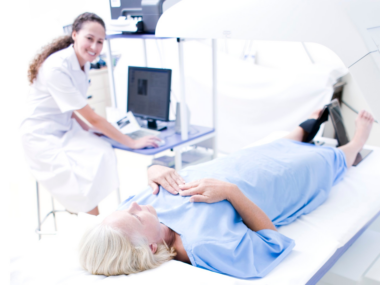 A photograph of a woman undergoing a DEXA bone density scan while a technologist operates an imaging machine